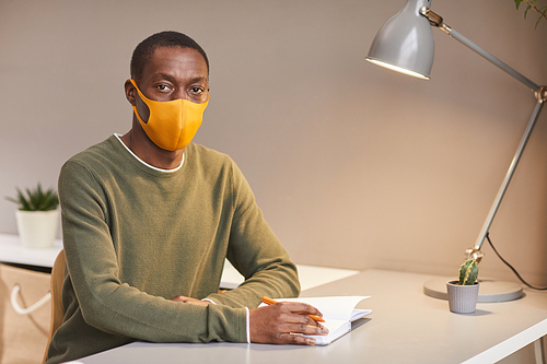 Portrait of African-American man wearing face mask and looking at camera while sitting at desk in home office, copy space