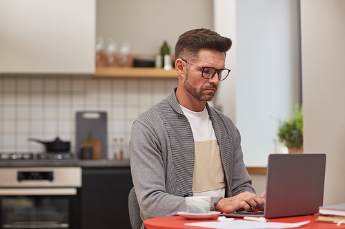 Side view portrait of handsome mature man working with laptop at home in kitchen interior, copy space