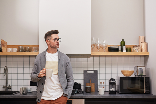 Waist up portrait of elegant adult man holding coffee mug and looking away pensively while relaxing standing in kitchen interior, copy space