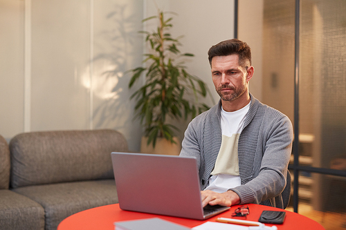 Portrait of handsome mature man working with laptop at red table in minimal office interior, copy space