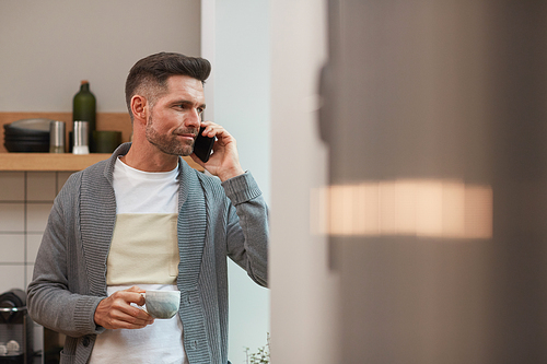 Waist up portrait of handsome mature man speaking by smartphone and enjoying cup of coffee in morning while standing by window in kitchen, copy space