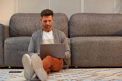 Full length portrait of handsome mature man working with laptop while sitting on floor at home in modern interior, copy space