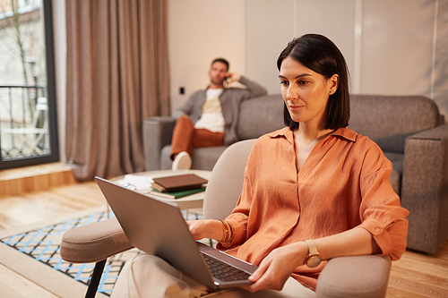 Portrait of modern couple working from home in minimal interior, focus on adult business woman using laptop while sitting in ergonomic armchair