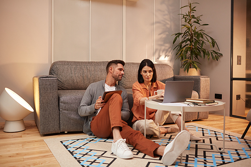 Full length portrait of modern adult couple working from home together while sitting on floor in stylish interior and using laptop, copy space