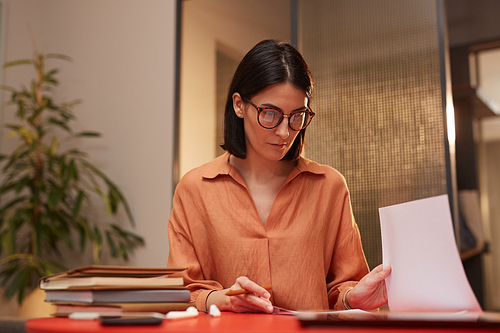 Portrait of modern adult businesswoman wearing glasses and sorting documents while working at desk in office lit by warm evening light, copy space