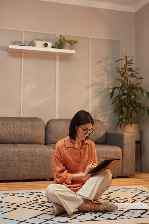 Full length portrait of contemporary woman working from home while sitting cross legged on floor and using digital tablet