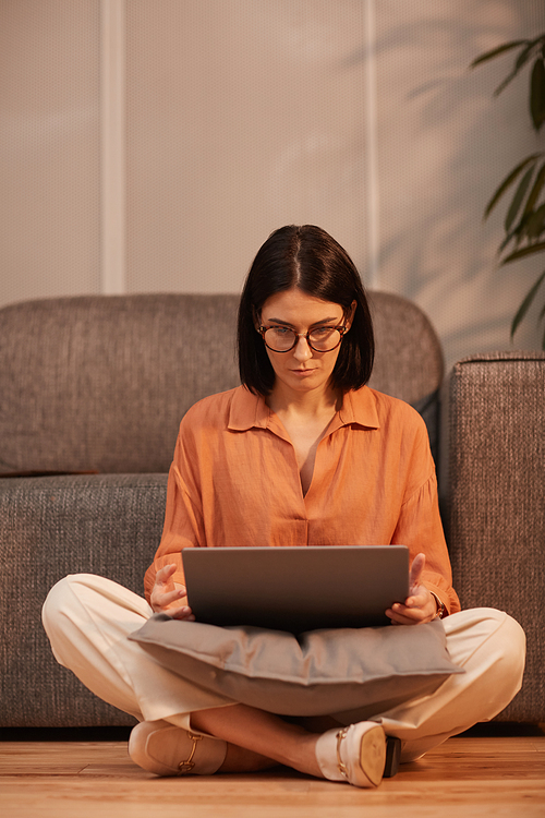 Full length portrait of contemporary woman working from home while sitting cross legged on floor and using laptop