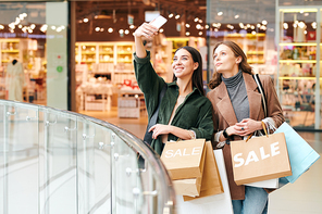 Young cheerful woman with smartphone and paperbags making selfie with friend standing next to her on background of mall interior