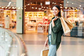 Portrait of cheerful young girl in green shirt standing paper bags with purchases in shopping mall