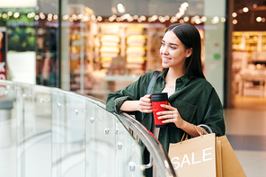 Smiling pretty brunette girl standing at railing and drinking hot tea from thermos cup in hall of shopping mall