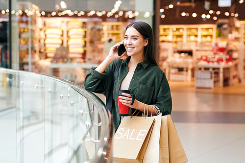 Young successful female customer with drink and paperbags talking on mobile phone after visiting departments of large modern mall