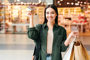 Young cheerful woman with paperbags and credit card looking at you with smile while expressing satisfaction with excellent shopping