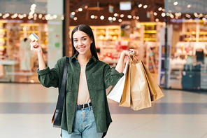 Portrait of excited young woman with paper bags using credit card during shopping in mall