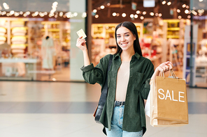 Young cheerful shopper with paperbags showing her credit card and looking at you while standing on background of mall departments