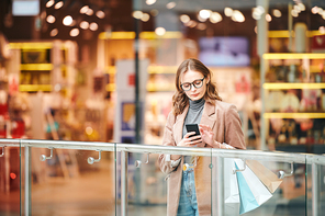 Serious young woman in eyeglasses standing at railing and checking smartphone in mall