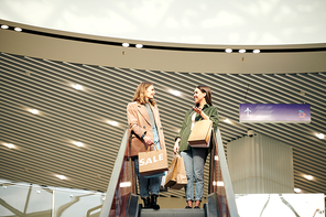 Two young women in casualwear holding paperbags while standing on elevator and discussing what they bought on sale in the mall