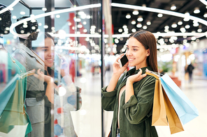Smiling student girl with paper bags standing at display case and looking at dress while asking advice of friend by phone