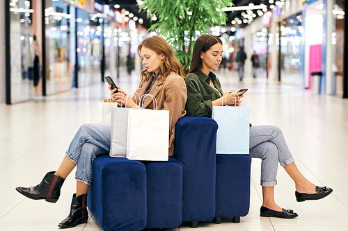 Serious young women sitting on poufs in mall lobby and using smartphones while searching for shopping special offers