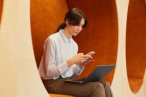 Portrait of modern young businesswoman using laptop and smartphone while working in open space office, sitting in graphic lounge zone, copy space