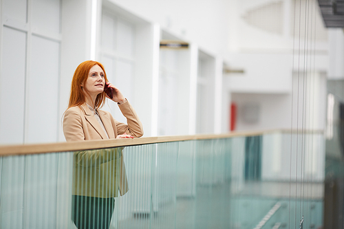 Portrait of mature red haired businesswoman speaking by smartphone and looking away while standing on balcony in white office building interior, copy space