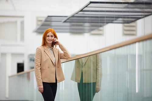 Portrait of mature red haired businesswoman speaking by smartphone and looking at camera while standing on balcony in white office building interior, copy space