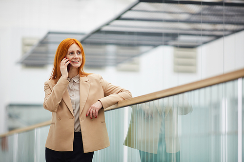 Waist up portrait of smiling red haired businesswoman speaking by smartphone and looking away while standing on balcony in white office building interior, copy space