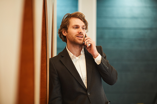 Waist up portrait of handsome bearded businessman speaking by smartphone and looking away while leaning on wall in minimal office interior, copy space