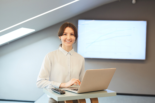 Waist up portrait of smiling young businesswoman looking at camera while posing at standing table with laptop in minimal interior with presentation screen in background, copy space