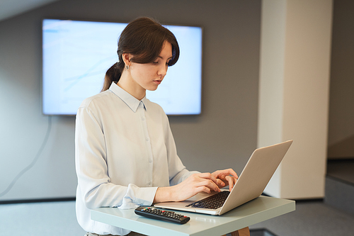 Waist up portrait of young businesswoman typing on laptop keyboard while working at standing table in minimal grey office interior, copy space