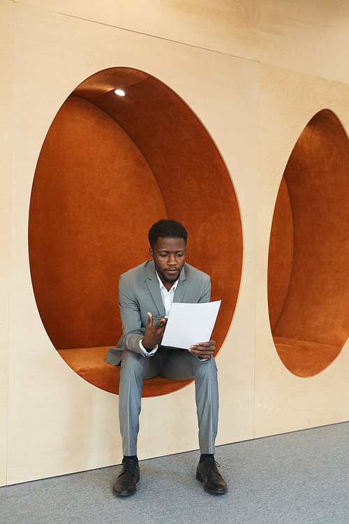 Vertical full length portrait of young African-American businessman reading documents while sitting in egg-shaped orange lounge zone of modern office or coworking space