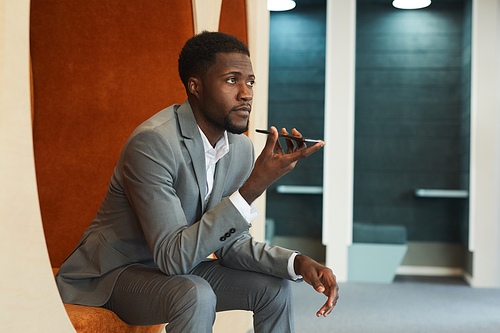 Portrait of handsome African-American businessman recording voice message via smartphone while sitting in lounge zone of modern office or coworking space