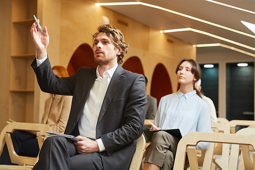 Portrait of successful businessman raising hand to ask question while sitting in audience of contemporary conference hall interior, copy space