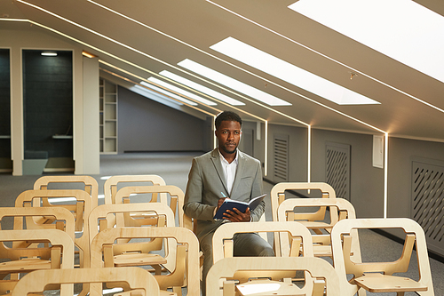 Portrait of successful African-American businessman writing in note pad and looking at camera while sitting in audience of empty conference hall interior, copy space