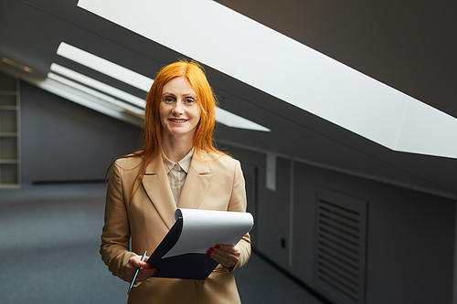 Waist up portrait of mature red-haired businesswoman holding clipboard and smiling at camera while standing in minimal office interior lit by skylights, copy space