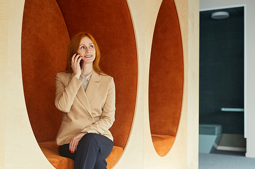 Portrait of mature red-haired businesswoman speaking by smartphone and smiling while sitting in lounge zone of modern office or coworking space
