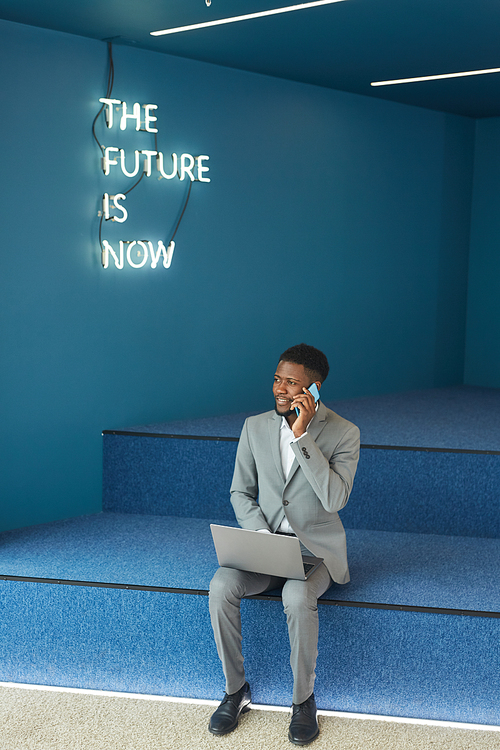 Vertical full length portrait of smiling African-American businessman using laptop and speaking by phone while working in blue lounge zone of modern office, copy space