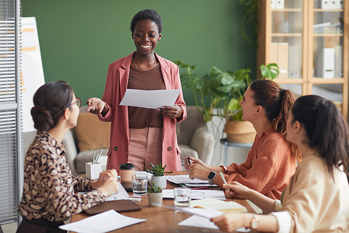 Portrait of successful African-American businesswoman holding documents while heading meeting with female team in office, copy space