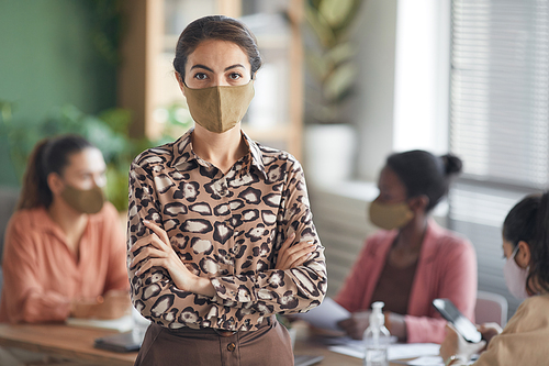 Waist up portrait of elegant businesswoman wearing mask and looking at camera while standing with arms crossed against business meeting in office, copy space