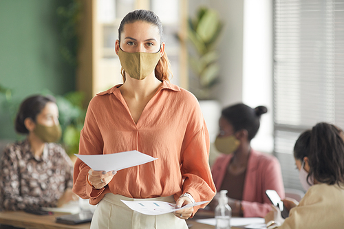 Waist up portrait of modern businesswoman wearing mask and looking at camera while standing against business meeting in office, copy space