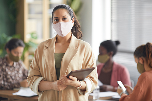 Waist up portrait of modern mixed-race businesswoman wearing mask and looking at camera while standing against business meeting in office, copy space