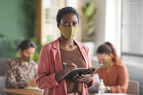 Waist up portrait of modern African-American businesswoman wearing mask and looking at camera while standing against business meeting in office, copy space