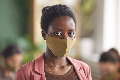 Portrait of modern African-American businesswoman wearing mask and looking at camera while standing against business meeting in office, copy space