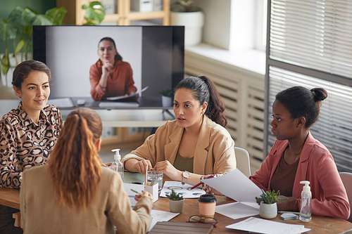 High angle view at multi-ethnic group of businesswomen discussing project during meeting in office with remote participant in background, copy space