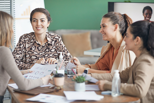 Multi-ethnic group of smiling businesswomen discussing project during meeting in office with remote participant in background, copy space