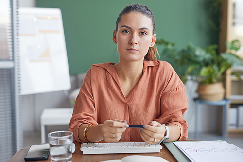 Portrait of contemporary businesswoman looking at camera with serious face expression while sitting at table in office against green wall