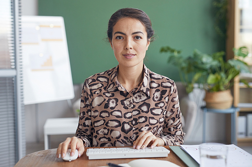 POV of elegant businesswoman looking at camera and using computer while sitting at table in office against green wall