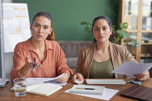 POV of two businesswomen looking at camera and pointing while using computer sitting at table in office against green wall