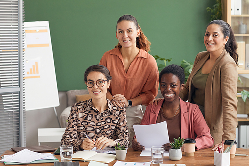 Multi-ethnic group of four contemporary businesswomen smiling at camera while posing in office, copy space