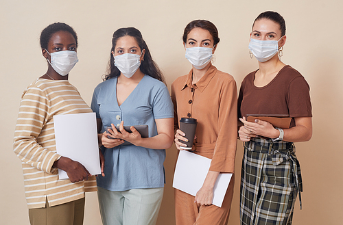 Multi-ethnic group of four young businesswomen wearing masks and looking at camera while standing by wall in office