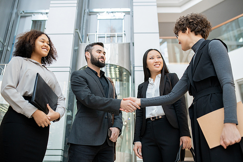 Curly manager with folder shaking hand of man while meeting with business team in office